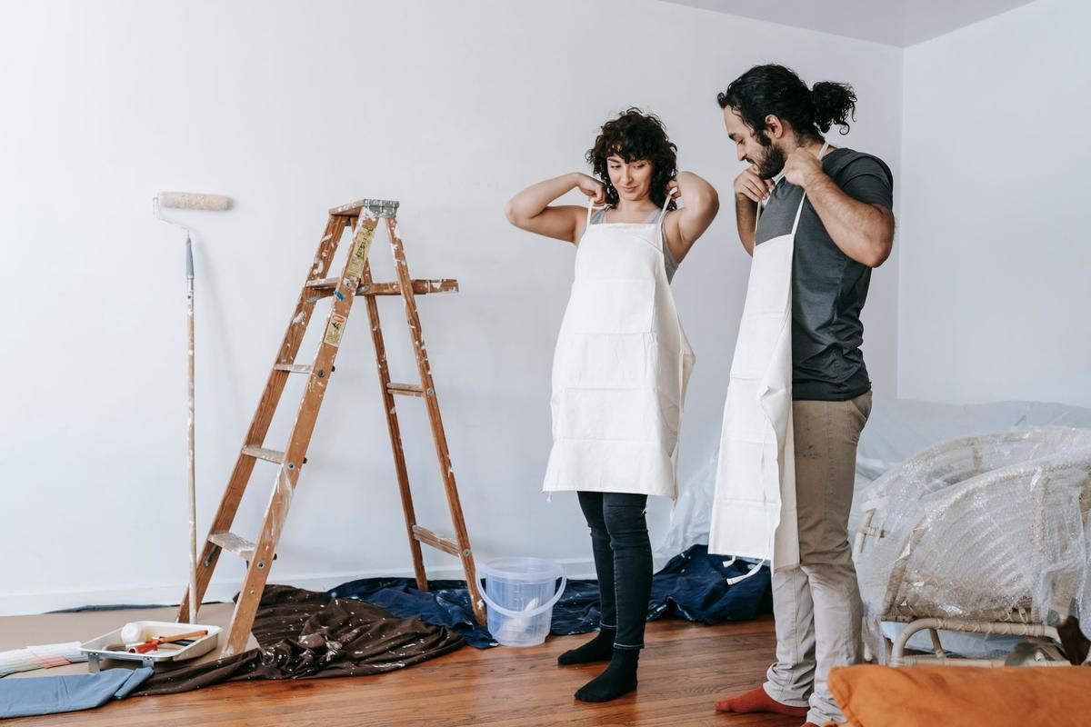 Deux personnes en tabliers blancs discutent devant un mur blanc avec échelle et matériel de peinture Deux personnes en tabliers blancs discutent devant un mur blanc avec échelle et matériel de peinture
